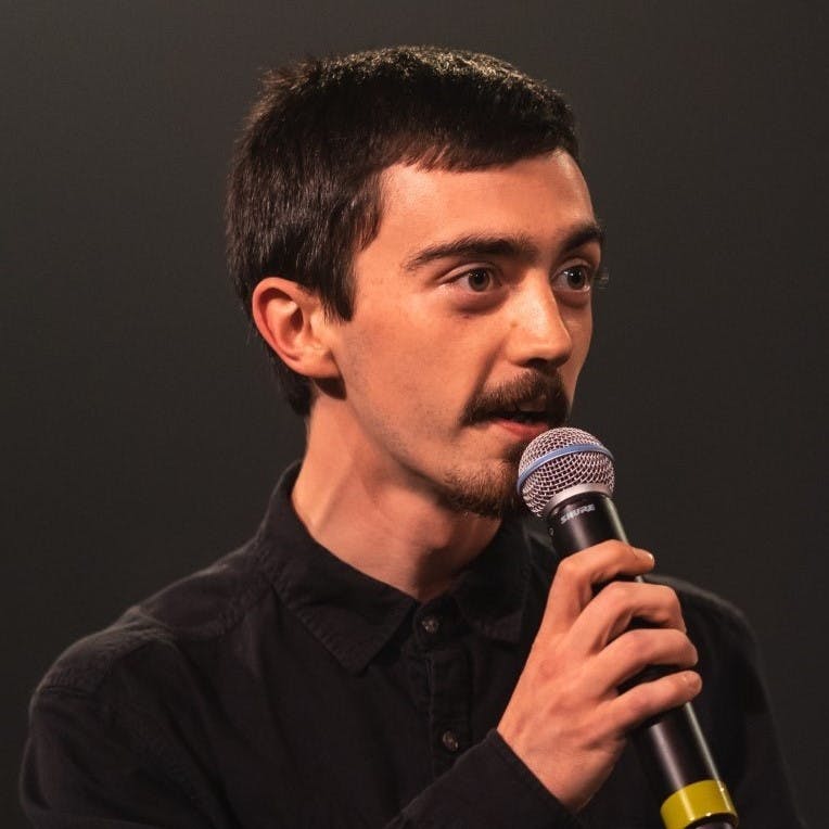 Cal hosting a performance. He is wearing a nice black shirt and has a microphone with a small piece of tape at the bottom of it in one hand. With the other hand, he seems to be describing something or telling a story to his audience who are not pictured.